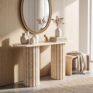 Può includere: Un tavolo console con un piano ovale liscio e due basi cilindriche scanalate in pietra beige chiaro. Il tavolo sostiene tre piccoli vasi in ceramica, una pila di libri e un piccolo oggetto dorato. Uno specchio ovale è appeso alla parete.