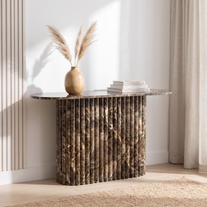 Op de afbeelding: Een ovale consoletafel met een donkerbruin marmeren blad en een geribbelde, cilindrische voet met een bijpassende marmeren afwerking. Een beige keramische vaas met gedroogd pampasgras en drie witte boeken staan op het tafeloppervlak.