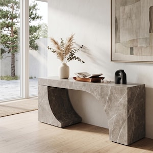 Op de afbeelding: Een grijze marmeren sidetable met een gebogen boogvoet. Bovenop staat een beige keramische vaas gevuld met gedroogd pampasgras en eucalyptus, een bruin lederen dienblad met een open boek, een bril en een zwart abstract sculptuur.