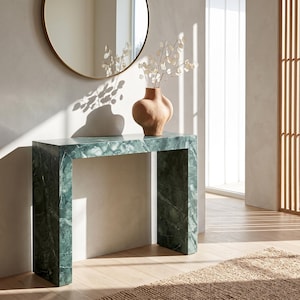 Puede incluir: Una mesa consola rectangular hecha de mármol verde con vetas blancas prominentes. La mesa presenta una silueta limpia y en forma de bloque. Un jarrón de cerámica color terracota con una forma redondeada y orgánica se asienta encima con tallos secos.