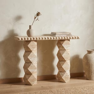 Puede incluir: Una mesa consola de travertino beige con un tablero rectangular de borde piramidal y dos columnas de soporte verticales compuestas por formas geométricas de diamante apiladas. Un pequeño jarrón y dos libros descansan sobre la superficie.