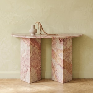 Può includere: Un tavolo console semicircolare realizzato in marmo venato rosa e bianco con due gambe a pilastro rettangolari. Un piccolo vaso in ceramica beige opaco poggia sulla superficie, contenente un singolo stelo botanico essiccato color marrone dorato.