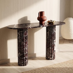 Può includere: Un tavolo console caratterizzato da un piano in marmo bordeaux scuro con venature bianche e due gambe cilindriche scanalate in marmo abbinato. Un vaso color terracotta opaco e una piccola ciotola dorata con pietre lisce poggiano sul piano.