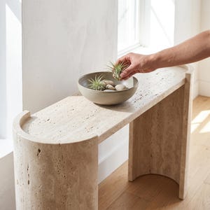 Peut inclure: Une console en travertin beige avec une base incurvée en demi-cercle et une surface supérieure lisse. Un bol en céramique ronde de couleur claire est posé sur la table, contenant des petits galets lisses et des plantes aériennes vertes.