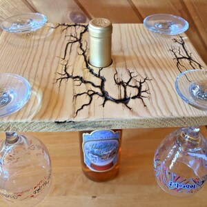 May include: A square light-colored wooden board with a central hole for a bottle neck and four corner cutouts for wine glasses. The wood features dark, branching fractal burn patterns. A glass bottle is inserted through the center hole.