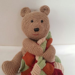 Puede incluir: Un oso de peluche tejido a mano de color marrón que sostiene una manta de retazos. El oso tiene pequeños ojos negros y una sonrisa roja cosida. La manta presenta cuadrados rojos, verdes, naranjas y beige, creando un ambiente acogedor y artesanal.