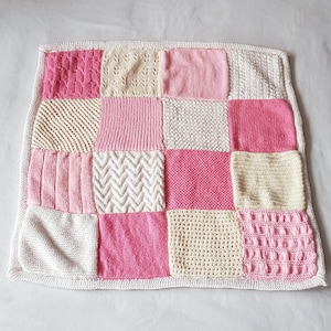 Puede incluir: Una manta de patchwork hecha a mano con una variedad de texturas y patrones de punto. La manta está compuesta por cuadrados en tonos de rosa, crema y blanco, con un borde blanco. Las diferentes texturas incluyen trenzas, canalé y punto de arroz.