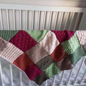 May include: A multi-coloured knitted baby blanket with a patchwork design. The blanket is made of different shades of green, red, brown, and pink.