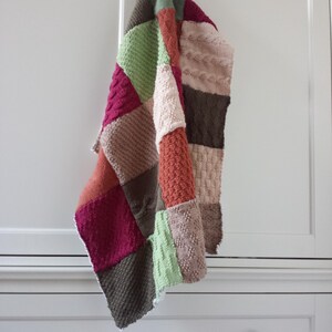 May include: A multi-coloured knitted patchwork blanket with various textures and patterns, including cable knit and plain knit. The blanket is hanging on a white cabinet door.