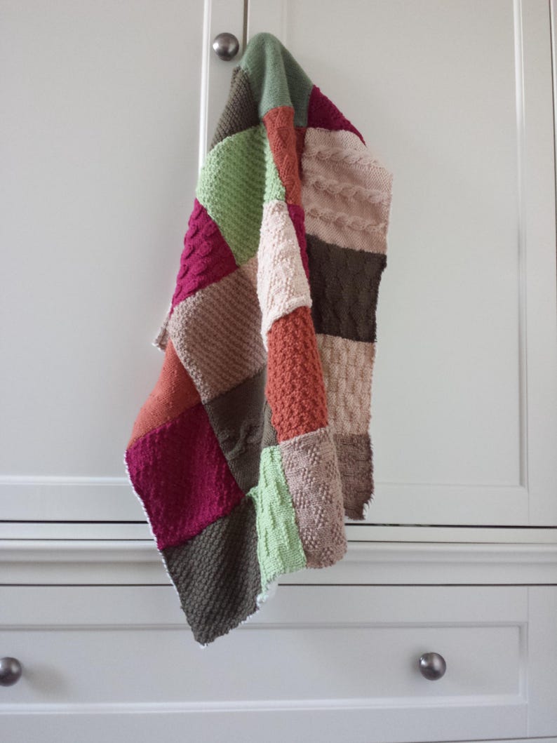 May include: A multi-coloured knitted patchwork blanket with various textures and patterns, including cable knit and plain knit. The blanket is hanging on a white cabinet door.
