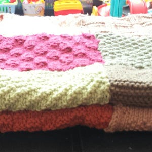 May include: A knitted patchwork blanket with squares in shades of pink, green, brown, and beige. The blanket is folded in half and has a soft, textured surface.