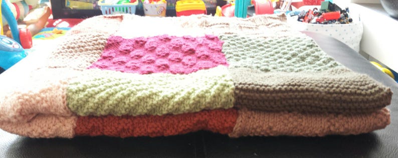 May include: A knitted patchwork blanket with squares in shades of pink, green, brown, and beige. The blanket is folded in half and has a soft, textured surface.
