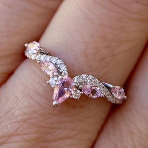 May include: A silver ring with a curved design, featuring pink gemstones and small clear stones. The ring has a delicate, elegant appearance, with the pink stones arranged in a floral or leaf-like pattern.