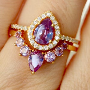 May include: A gold ring set with a pear-shaped purple gemstone surrounded by smaller clear gemstones. A second band with smaller purple gemstones is worn below the main ring.