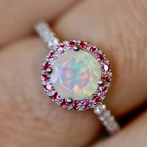 May include: A close-up of a silver ring featuring a large, round opal gemstone surrounded by a halo of small, pink gemstones. The ring's band is also adorned with smaller, clear gemstones, creating a sparkling effect. The opal has a play of color.