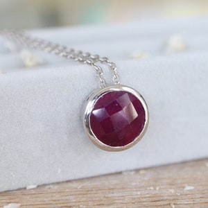 May include: A silver chain necklace with a round pendant featuring a deep red ruby gemstone.