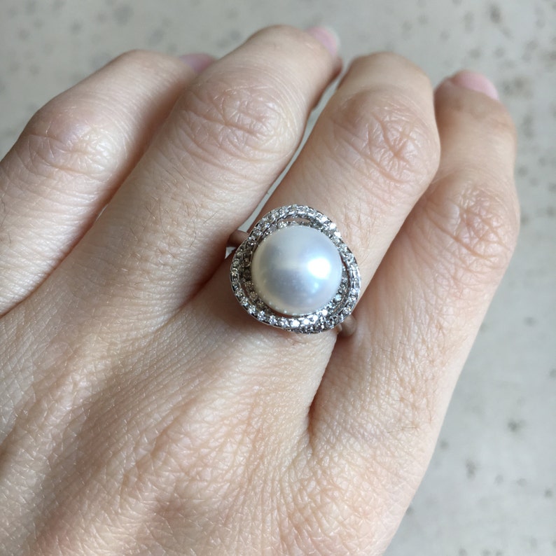 May include: A silver ring with a large white pearl surrounded by a halo of smaller clear stones.