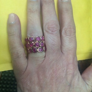 May include: A gold ring with multiple rows of oval-shaped pink gemstones. The ring is worn on a person's finger.