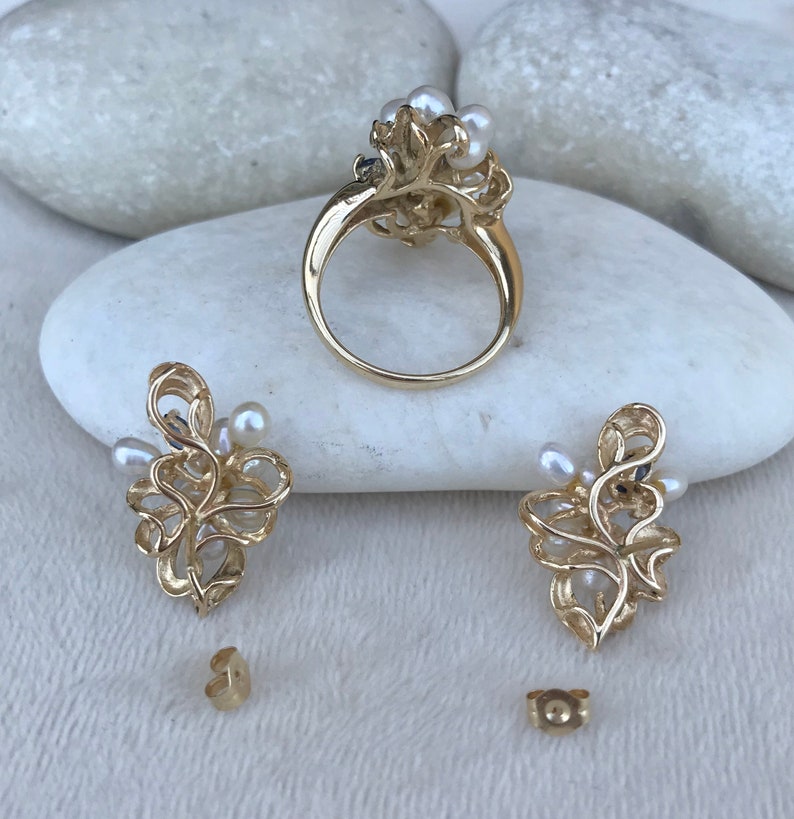 May include: A gold ring and earrings set with a delicate floral design and white pearls. The ring has a wide band and the earrings are drop style.