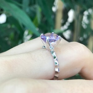 May include: A silver ring with a large, light purple gemstone set in a prong setting. The band is textured and has smaller green gemstones.