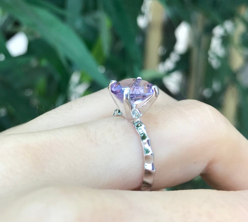 May include: A silver ring with a large, light purple gemstone set in a prong setting. The band is textured and has smaller green gemstones.