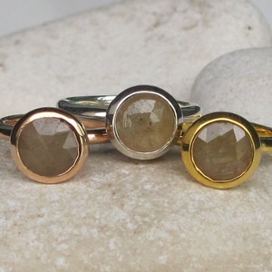 Op de afbeelding: Drie ringen met lichtbruine edelstenen, gezet in roségoud, zilver en goud. De gefacetteerde edelstenen zijn rond. De ringen worden gepresenteerd op een getextureerd, licht oppervlak.