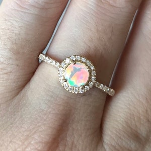 Pode incluir: Um anel de ouro com uma gema de opala redonda cercada por um halo de gemas menores e transparentes.