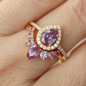 May include: A gold ring with a large pear-shaped purple gemstone surrounded by smaller clear gemstones. The ring is set with a smaller band of clear gemstones.