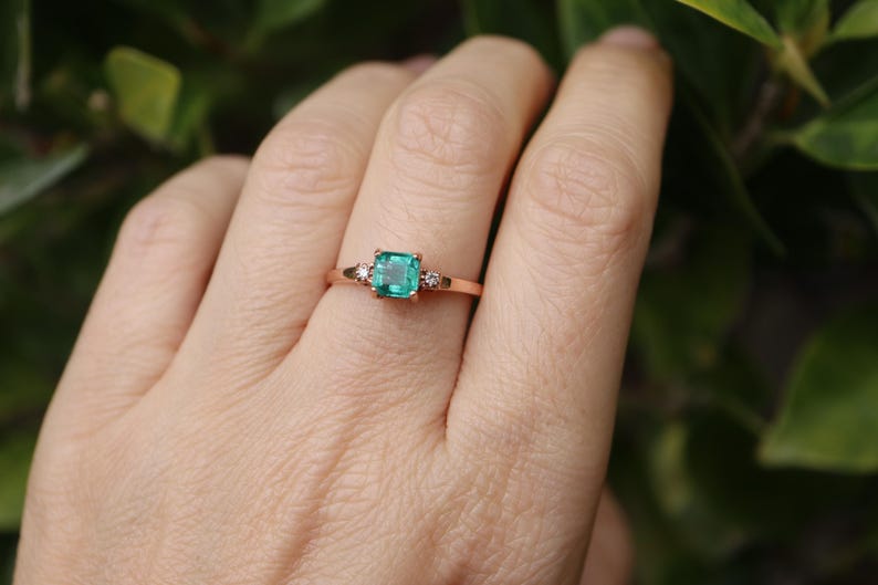 Op de afbeelding: Een ring van ros&eacute;goud met een vierkante smaragdgroene edelsteen en twee kleinere ronde, heldere edelstenen op de band.