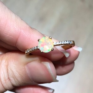 Pode incluir: Um anel de ouro com uma pedra de opala branca e uma banda de pequenas pedras transparentes.