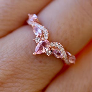 May include: A rose gold ring with a curved design, featuring pink pear-shaped gemstones and small, clear accent stones. The ring's gemstones are set in a delicate, floral-inspired pattern, creating a sparkling and elegant look.