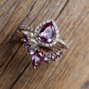 May include: Two silver rings with purple gemstones. The top ring features a large pear-shaped gemstone surrounded by smaller diamonds. The bottom ring has a row of smaller pear-shaped gemstones.