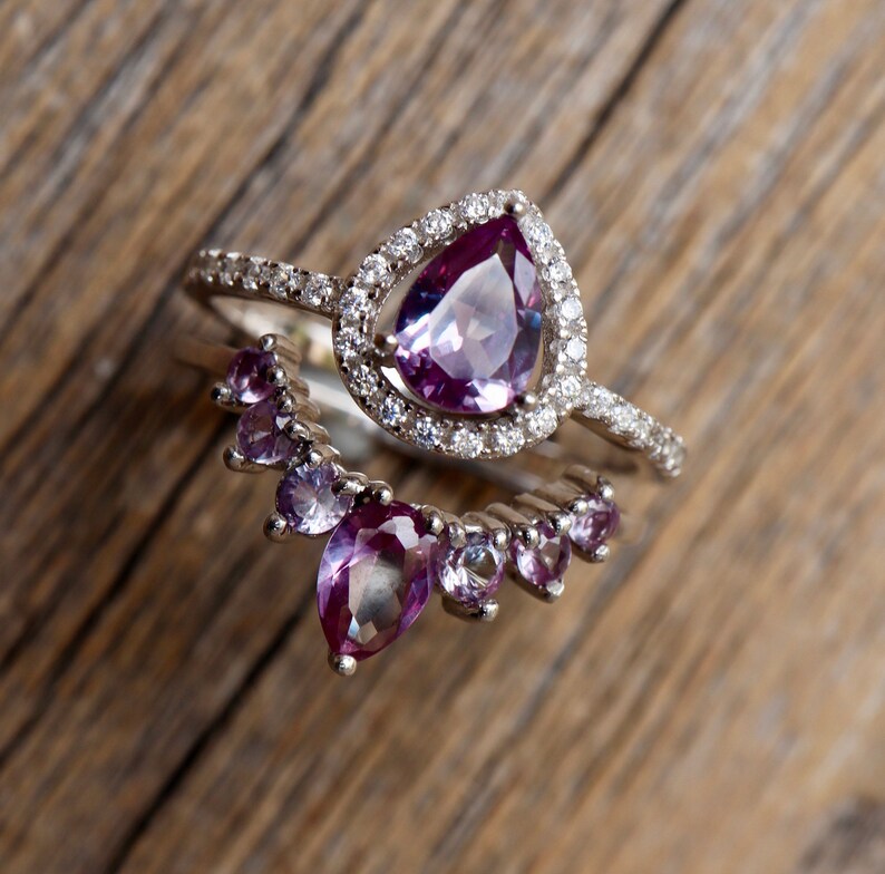 May include: Two silver rings with purple gemstones. The top ring features a large pear-shaped gemstone surrounded by smaller diamonds. The bottom ring has a row of smaller pear-shaped gemstones.