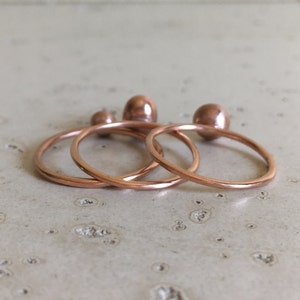 May include: A set of four copper-colored rings and three matching copper-colored spheres. The rings are thin and circular, and the spheres are small and smooth. The items are arranged on a light-colored surface.