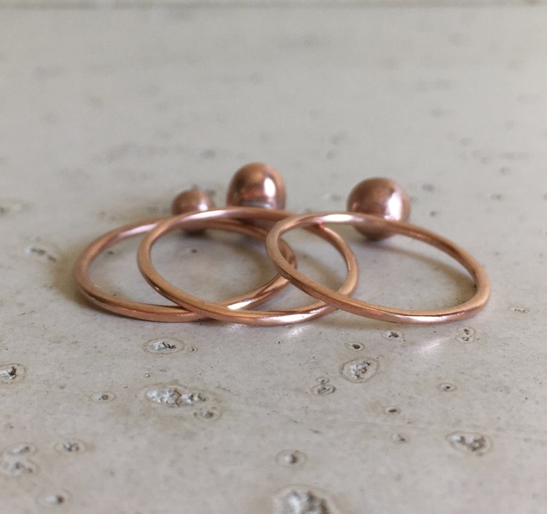 May include: A set of four copper-colored rings and three matching copper-colored spheres. The rings are thin and circular, and the spheres are small and smooth. The items are arranged on a light-colored surface.