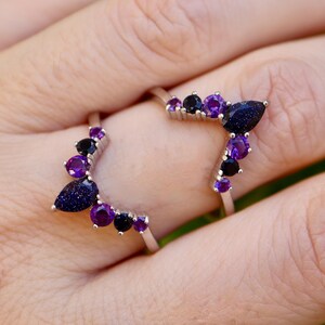 May include: Two silver rings, each adorned with a cluster of gemstones. The rings feature a dark blue, teardrop-shaped stone, complemented by purple and black gems. Set against a blurred green backdrop.