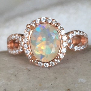 Pode incluir: Um anel de ouro rosa com uma grande pedra de opala ovalada cercada por um halo de pedras preciosas menores e transparentes. A banda também é decorada com pedras preciosas menores e transparentes.