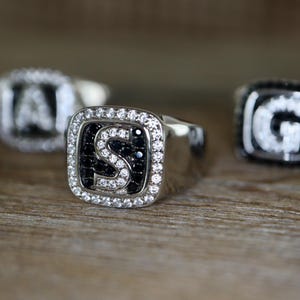 Peut inclure: Bagues en argent avec des faces carrées, chacune avec une lettre. La bague centrale affiche la lettre "S" en blanc, entourée de pierres noires et blanches. D'autres bagues avec les lettres "A" et "G" sont visibles.
