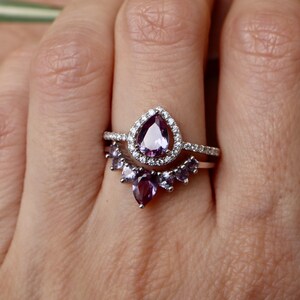 May include: A silver ring set with a pear-shaped purple gemstone surrounded by smaller clear gemstones. The ring is paired with a matching band featuring a row of smaller purple gemstones.