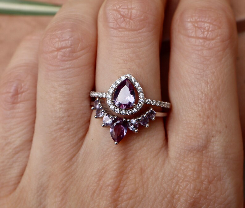 May include: A silver ring set with a pear-shaped purple gemstone surrounded by smaller clear gemstones. The ring is paired with a matching band featuring a row of smaller purple gemstones.