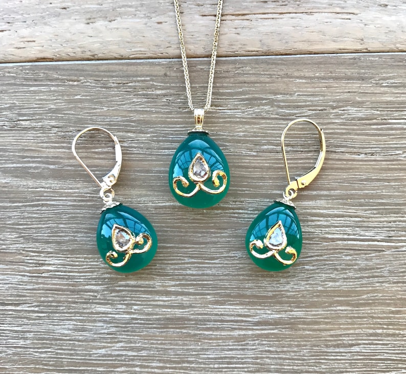 Op de afbeelding: Een groene edelsteen ketting met hanger en bijpassende oorbellen met gouden accenten en heldere strass steentjes. De traanvormige stenen zijn gezet in goud en hebben een delicaat bloemmotief.