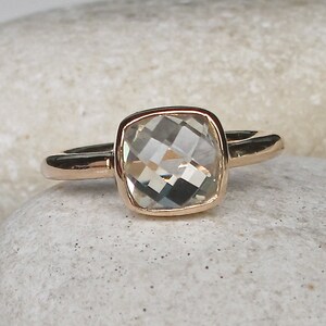 Puede incluir: Un anillo de oro con una piedra preciosa cuadrada transparente. El anillo est&aacute; sobre un fondo blanco.