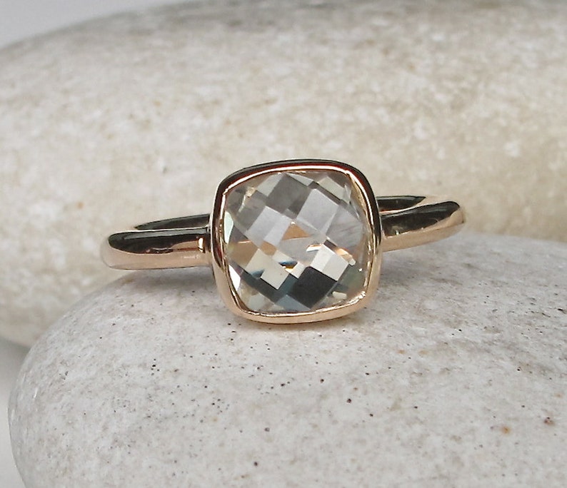 Puede incluir: Un anillo de oro con una piedra preciosa cuadrada transparente. El anillo est&aacute; sobre un fondo blanco.
