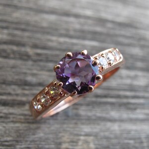 May include: A rose gold ring with a large, round, purple amethyst gemstone surrounded by smaller, clear gemstones.