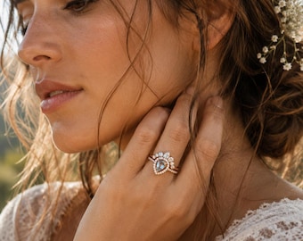 May include: A gold ring set featuring a central pear-shaped blue gemstone surrounded by a halo of small white stones, paired with a curved band adorned with marquise and round white stones, worn on a finger with a delicate metal finish.
