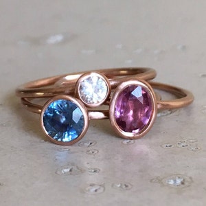 May include: Three rings with rose gold bands. One ring features a round, light blue gemstone, another a round, clear gemstone, and the third an oval, pink gemstone. The rings are displayed on a neutral surface.