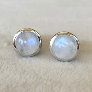 May include: A pair of round stud earrings featuring smooth, translucent moonstones with subtle blue iridescence, each set in a polished silver-toned metal bezel. The stones have a domed cabochon cut, reflecting light with a soft, milky glow.