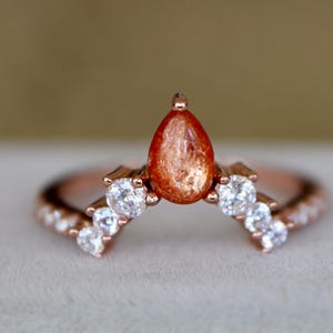 May include: Rose gold ring featuring a teardrop-shaped, orange gemstone at the center, flanked by three small, round, clear gemstones on each side. The band is adorned with tiny, clear gemstones. The ring is set against a soft, neutral background.