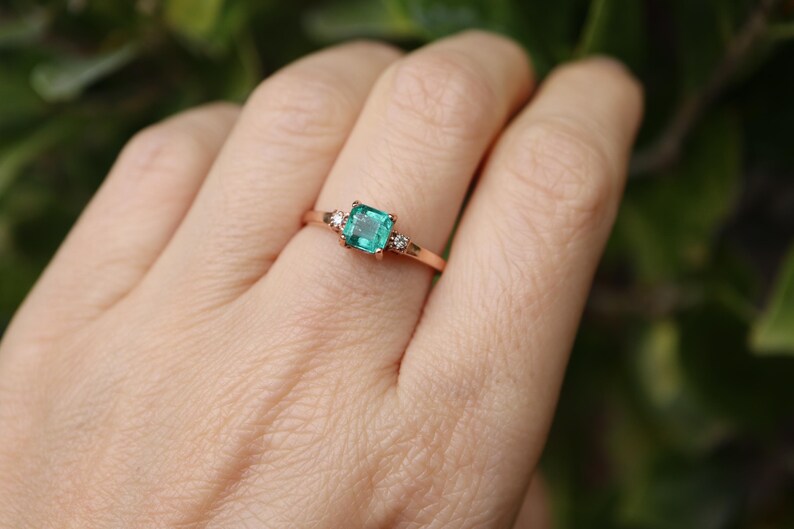 Op de afbeelding: Een ring van ros&eacute;goud met een vierkante smaragdgroene edelsteen en twee kleinere ronde diamanten aan weerszijden.