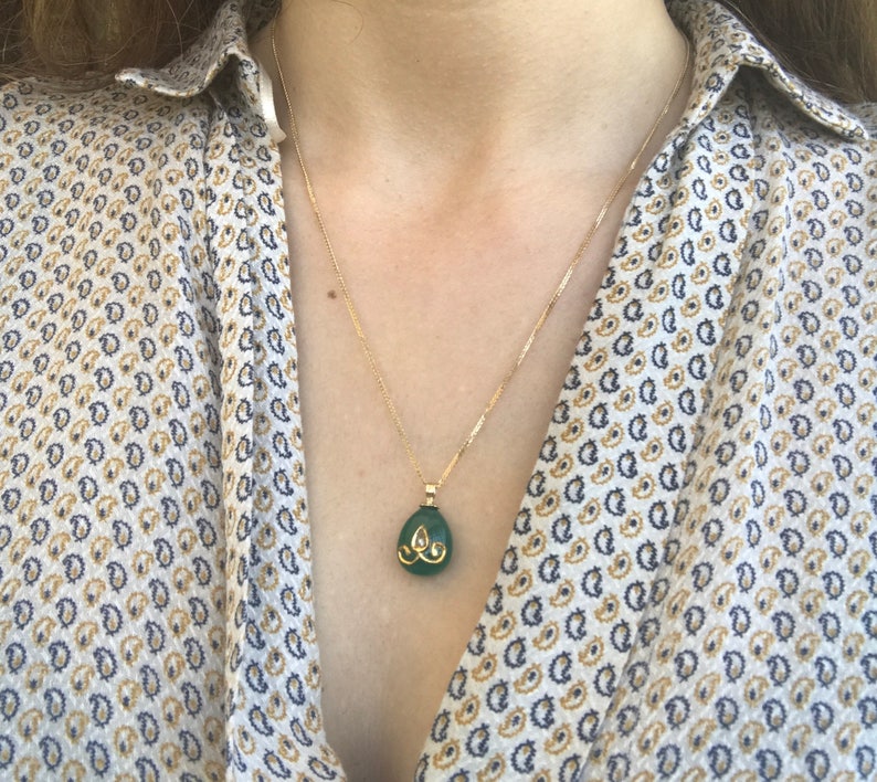 Op de afbeelding: Een gouden ketting met een groene edelsteen hanger. De hanger heeft de vorm van een traan en heeft gouden accenten.